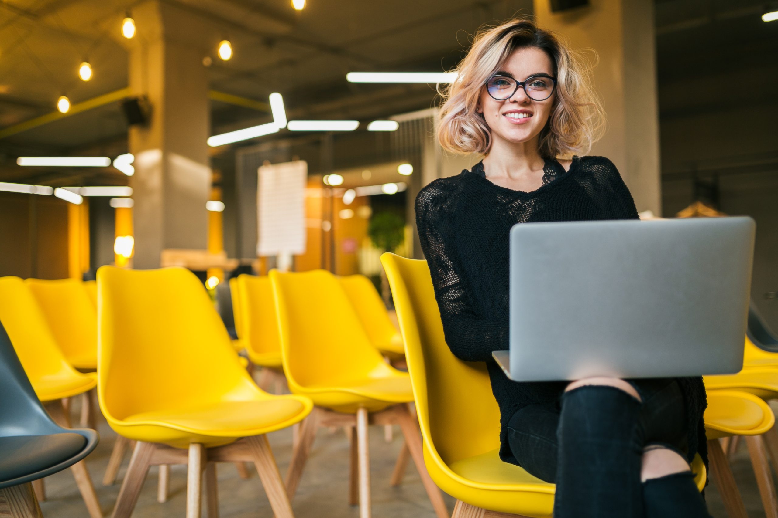 portrait of young attractive woman sitting in lecture hall, working on laptop, wearing glasses, classroom with many yellow chairs, student learning, education online, freelancer, smiling, stylish